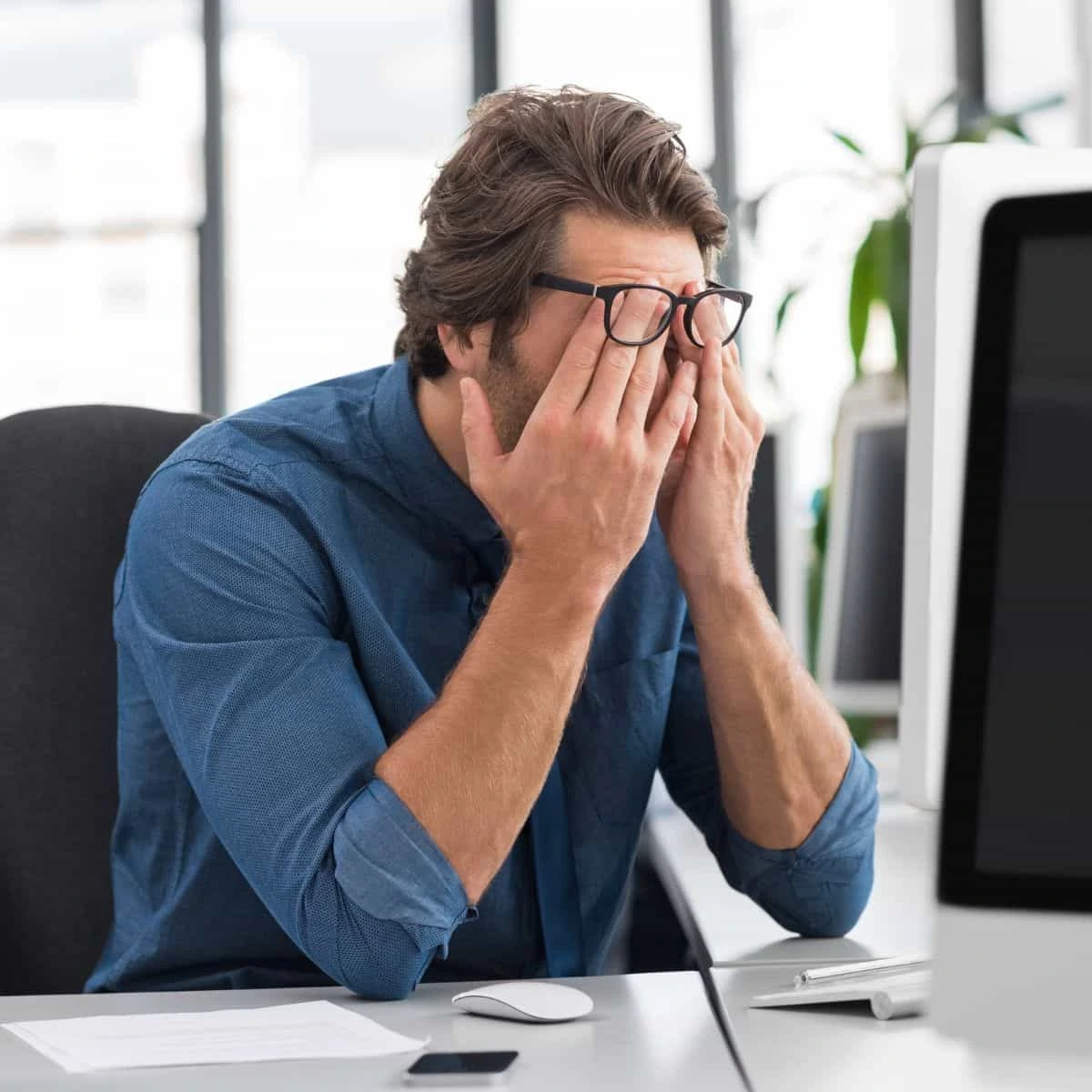 Un empleado de oficina sentado en su escritorio se cubre los ojos y el rostro con las manos en un gesto de severo agotamiento frente a la pantalla de su ordenador, ilustrando el software SaaS para la detección temprana y reducción del burnout corporativo y la fatiga laboral en entornos corporativos.